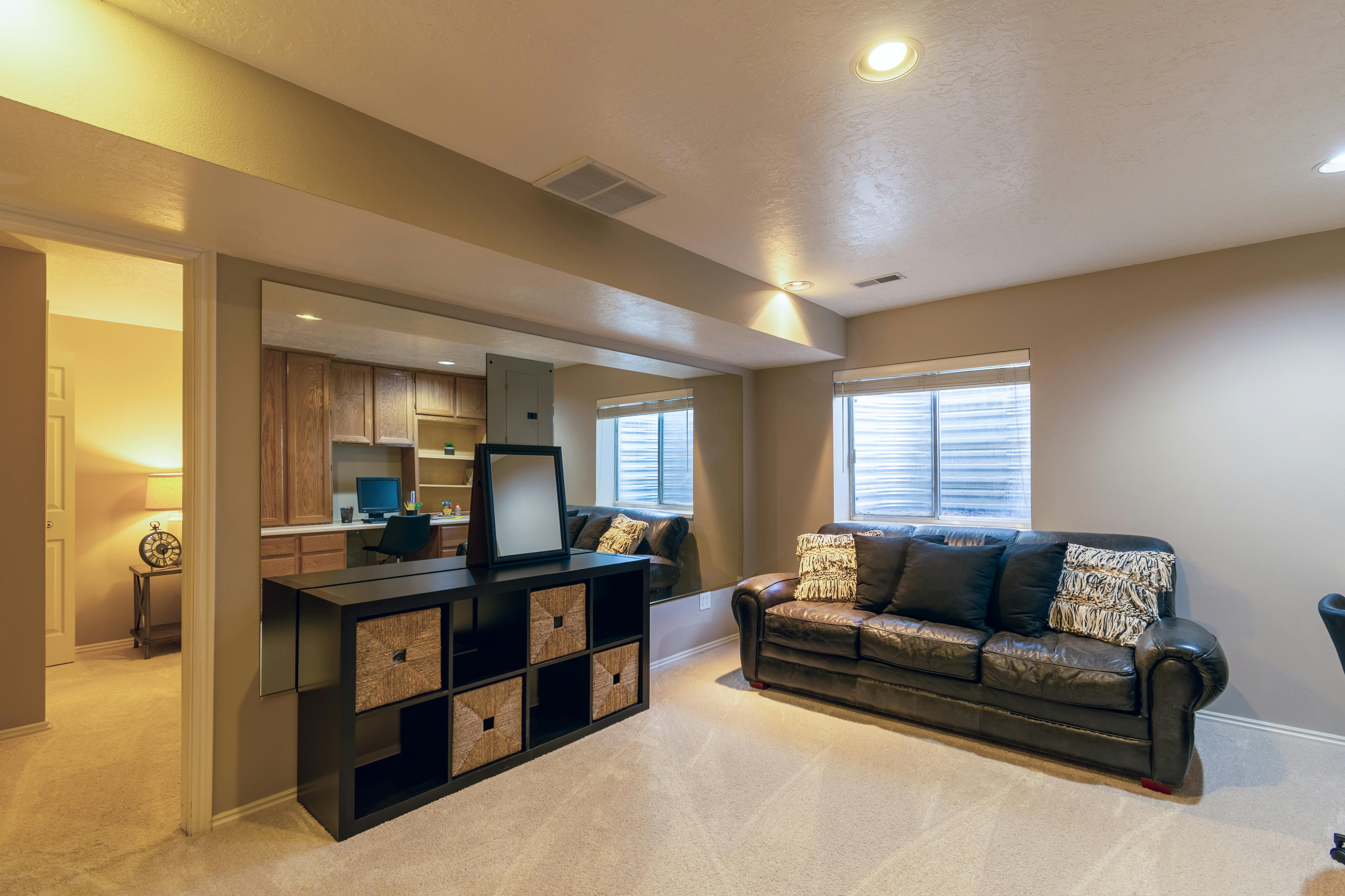 Finished basement with integrated home office, black leather couch, storage console, and natural light from egress windows.